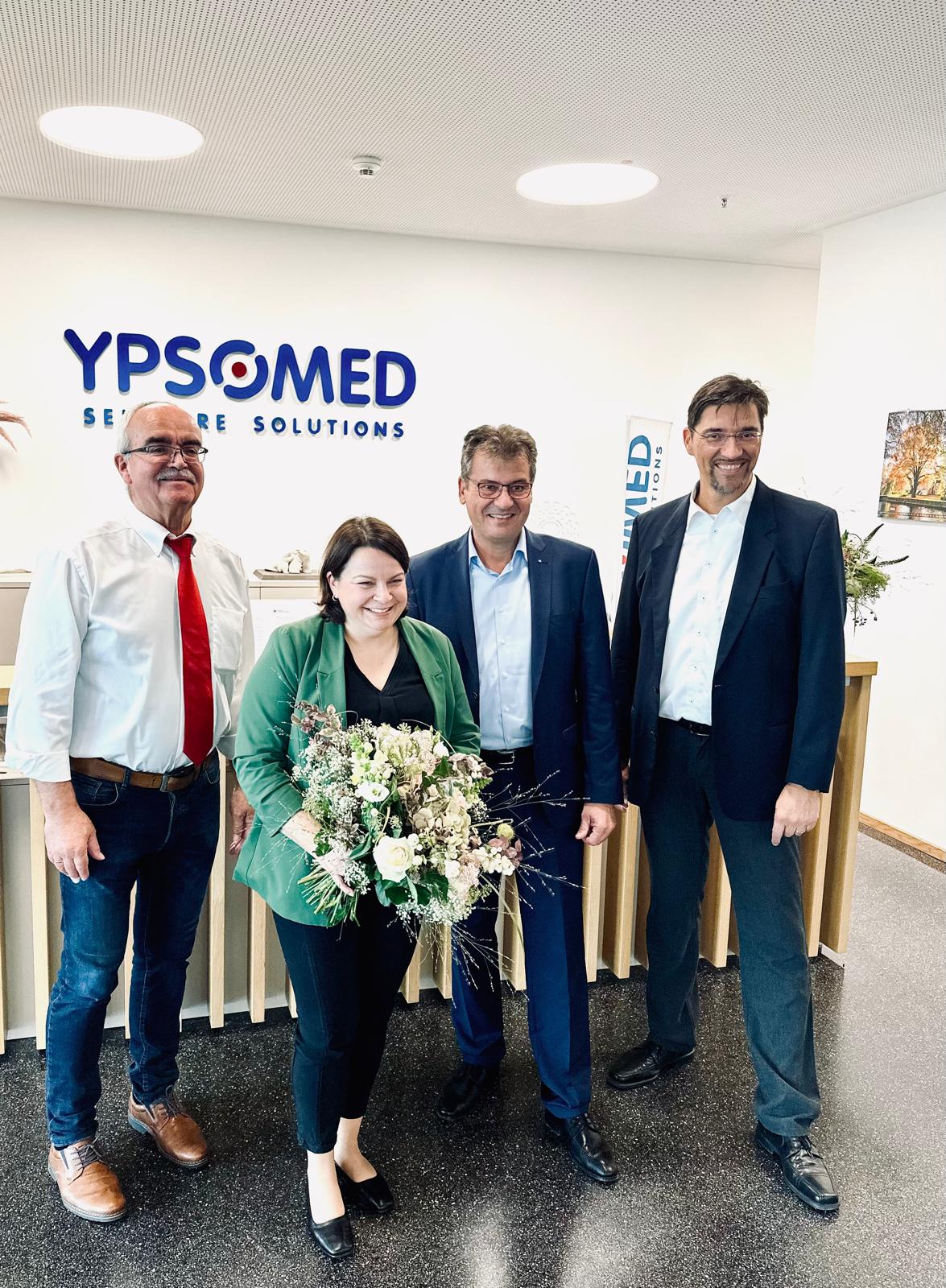 Vier Personen stehen lächelnd in einem Büro vor einem Ypsomed-Logo, eine Frau hält einen Blumenstrauß.