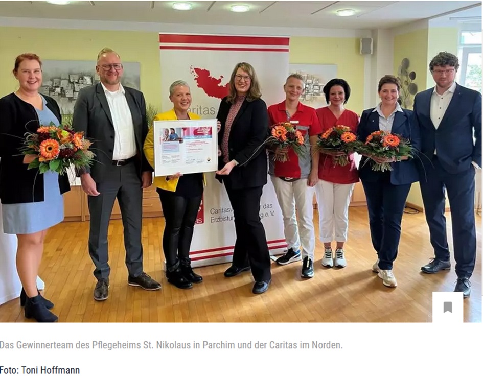 Gruppe von sieben Menschen, einige mit Blumen, lächeln vor einem Caritas-Banner. Eine hält eine Auszeichnung.