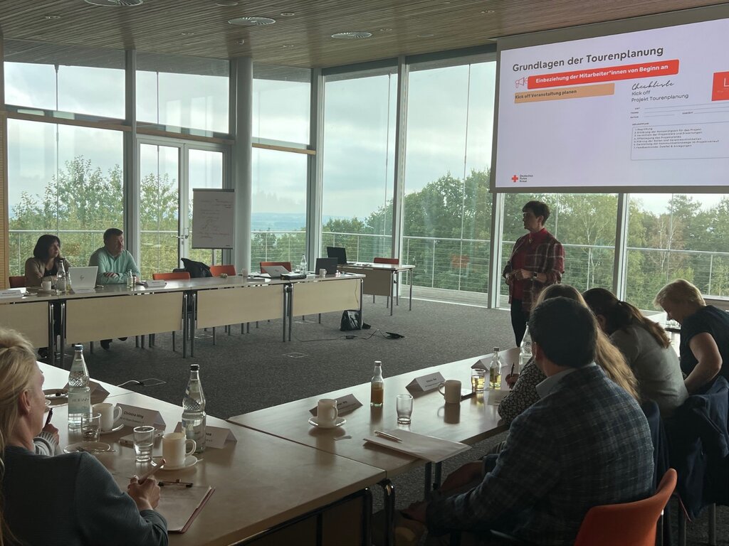 Person hält Vortrag in Konferenzraum mit Präsentation "Grundlagen der Tourenplanung", Teilnehmer sitzen an Tischen.
