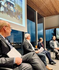 Vier Personen diskutieren auf einem Podium, Hintergrund zeigt ein Foto auf einer Leinwand.