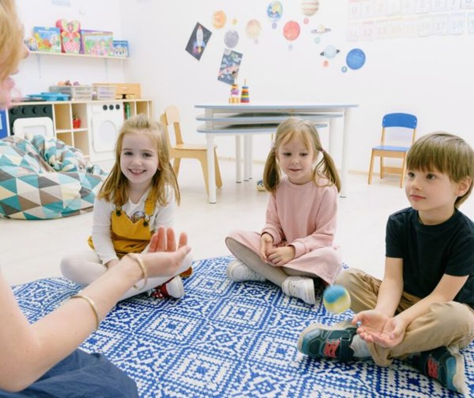Drei Kinder sitzen im Kreis auf einem Teppich, spielen mit einem Ball. Erwachsenenhand wirft den Ball. Wand mit Weltall-Deko.