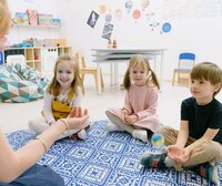 Drei Kinder sitzen im Kreis auf einem Teppich, spielen mit einem Ball. Erwachsenenhand wirft den Ball. Wand mit Weltall-Deko.