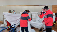 Personen in roten Jacken falten weiße Laken in einem Innenraum. Im Hintergrund stehen gestapelte Bettgestelle.