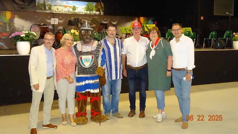 Ehrenamtmessen in Torgelow Sechs Personen und eine Person in bayerischem Trachtenkostüm mit Hut, Maske und Stock stehen in einer Halle vor einer Bühne mit Musikinstrumenten.