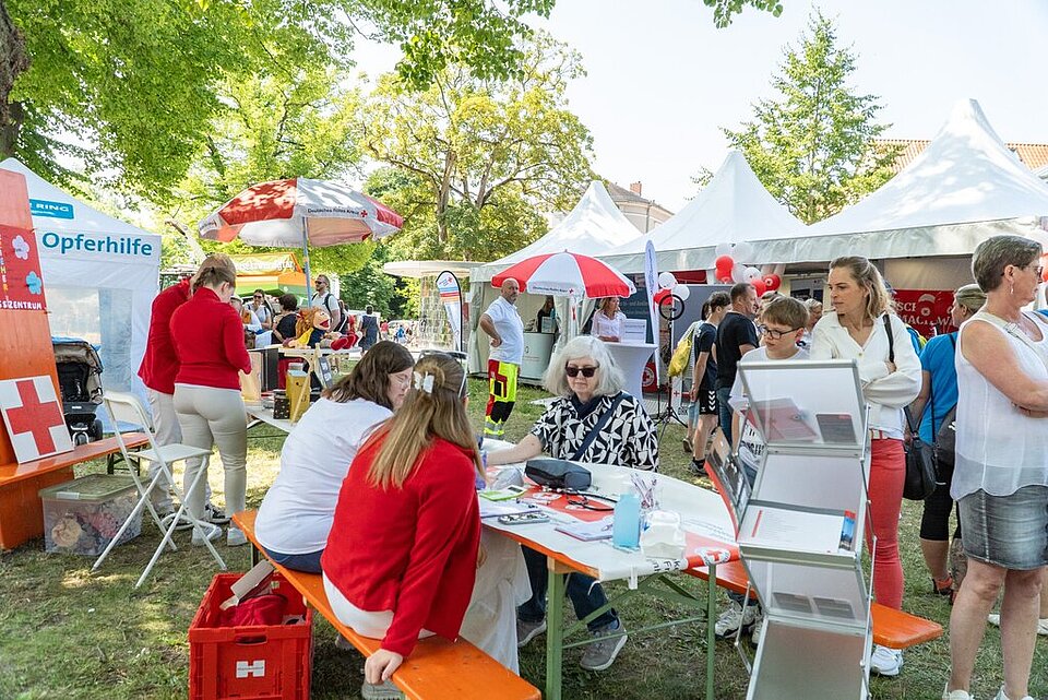 Menschen an Informationsständen im Freien bei einer Veranstaltung mit weißen Zelten und Bäumen im Hintergrund