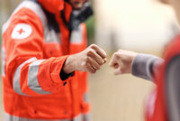 Zwei Personen geben sich einen Faustgruß, eine trägt eine orange Rettungsjacke mit Rotkreuz-Logo
