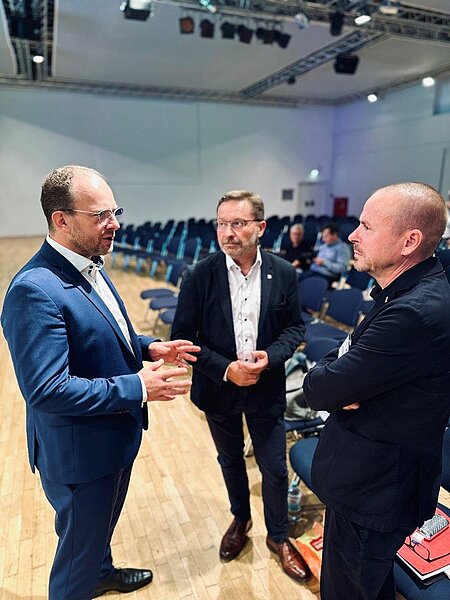 Drei Männer im Anzug unterhalten sich in einem Saal mit leeren Stuhlreihen im Hintergrund.