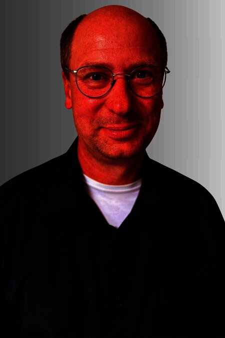 Mann mit Brille lächelt in die Kamera. Er trägt ein dunkles Hemd. Hintergrund ist hellgrau.