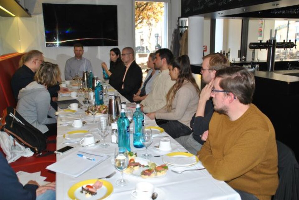Menschen sitzen an einem Tisch bei einem Meeting, mit Getränken und Snacks, in einem Raum mit Fernseher an der Wand.