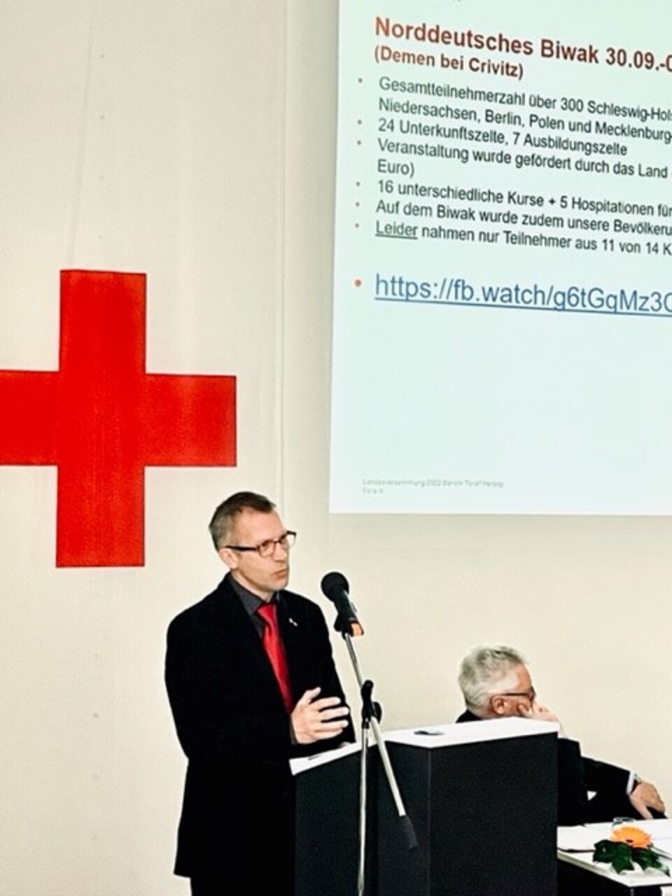 Mann hält Vortrag vor Leinwand mit Text. Großes rotes Kreuz im Hintergrund.