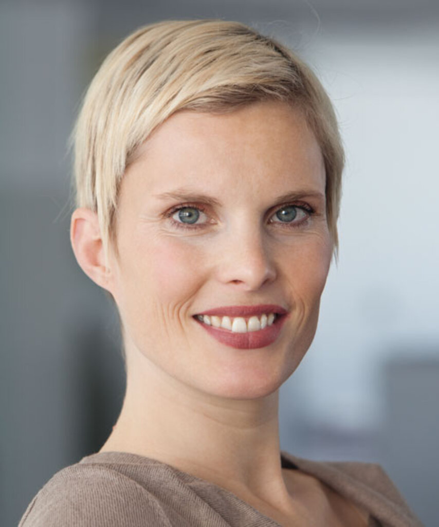 Lächelnde Frau mit kurzem blondem Haar vor unscharfem Hintergrund.