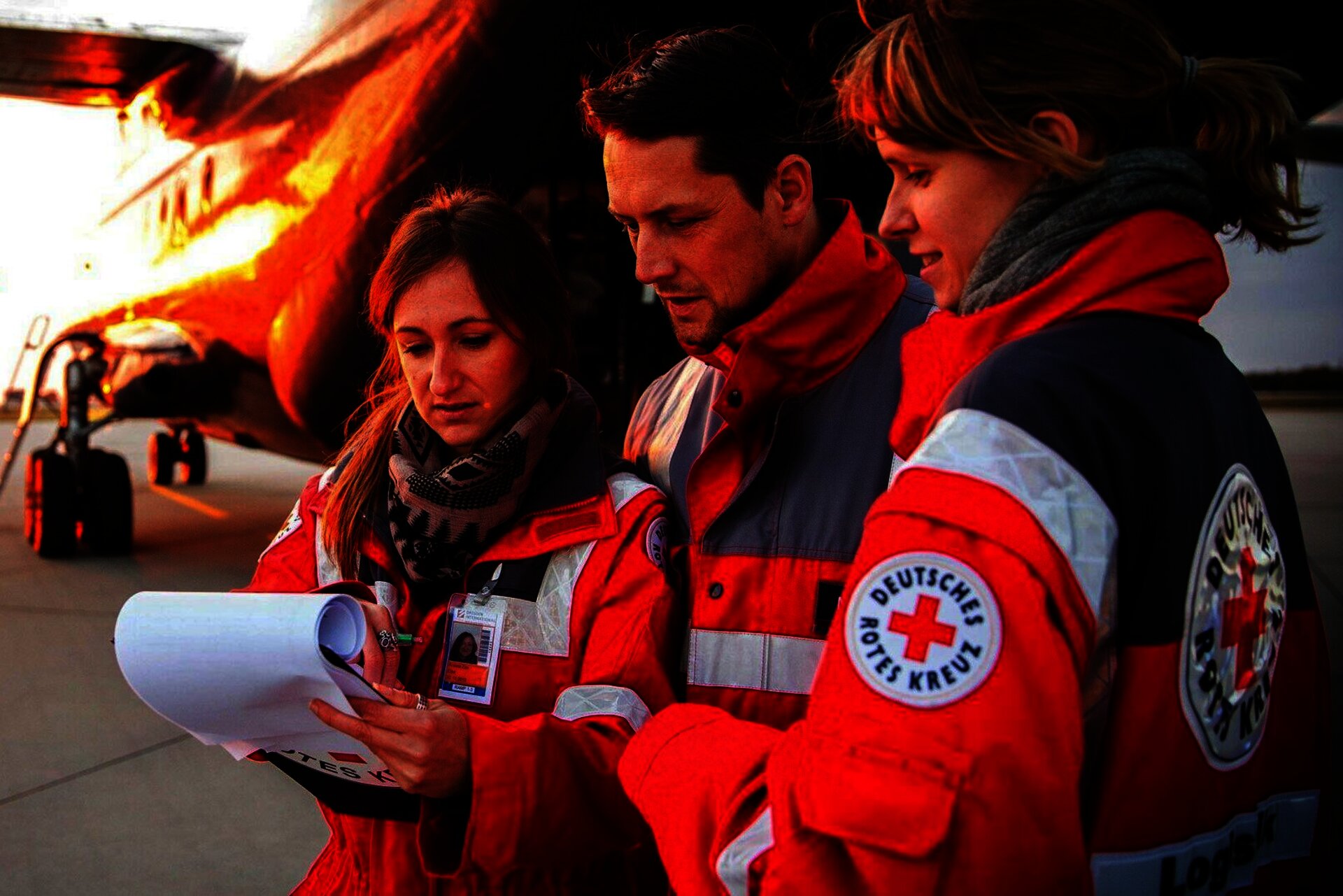 Drei Personen des Roten Kreuzes stehen vor einem Flugzeug und betrachten Dokumente bei Sonnenuntergang.