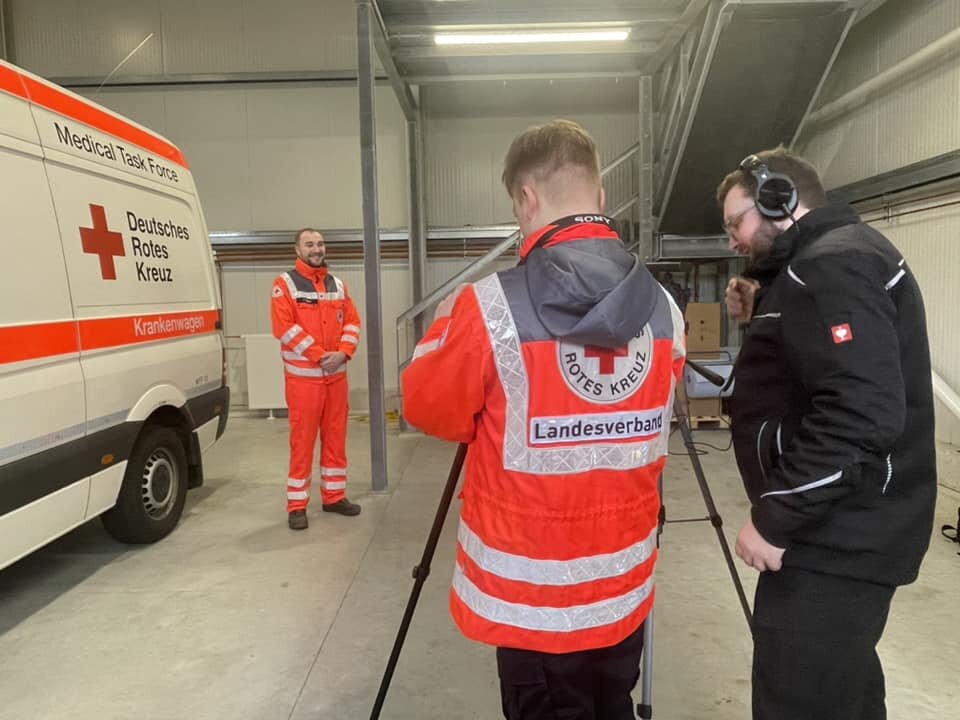Zwei Männer filmen einen Mann in DRK-Uniform neben einem Krankenwagen in einer Halle.