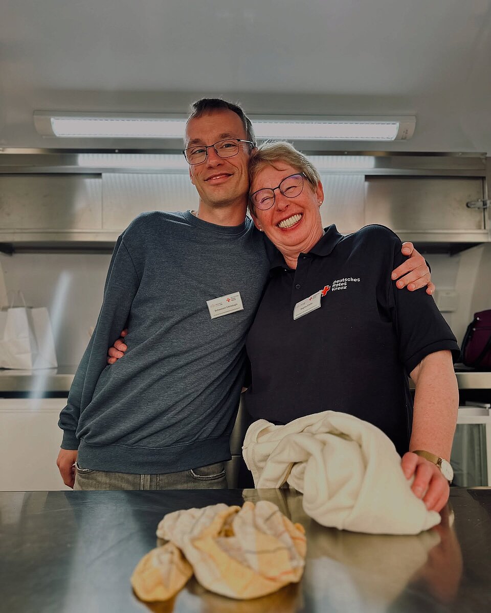 Zwei lächelnde Personen in Uniform umarmen sich in einer Küche, Küchentücher im Vordergrund.