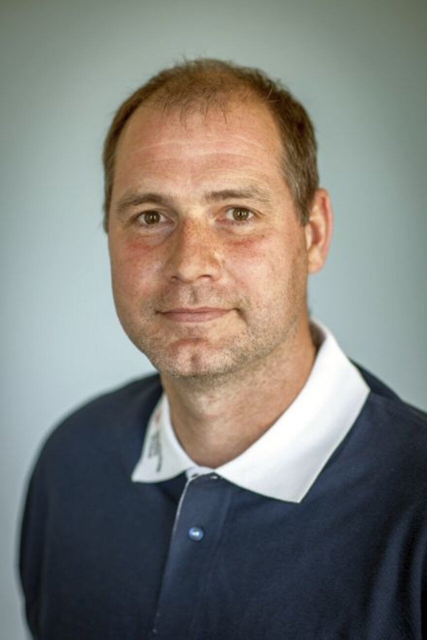 Mann mit blauem Hemd und weißem Kragen, schaut lächelnd in die Kamera, neutraler Hintergrund.