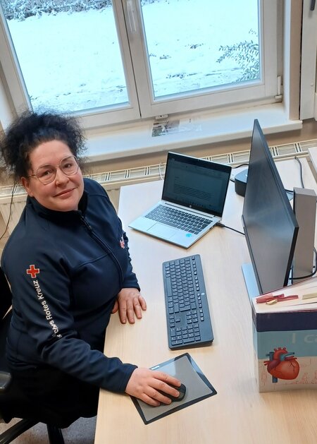 Frau im Rot-Kreuz-Pullover arbeitet am Schreibtisch mit Laptop und Monitor, winterliche Aussicht aus dem Fenster.