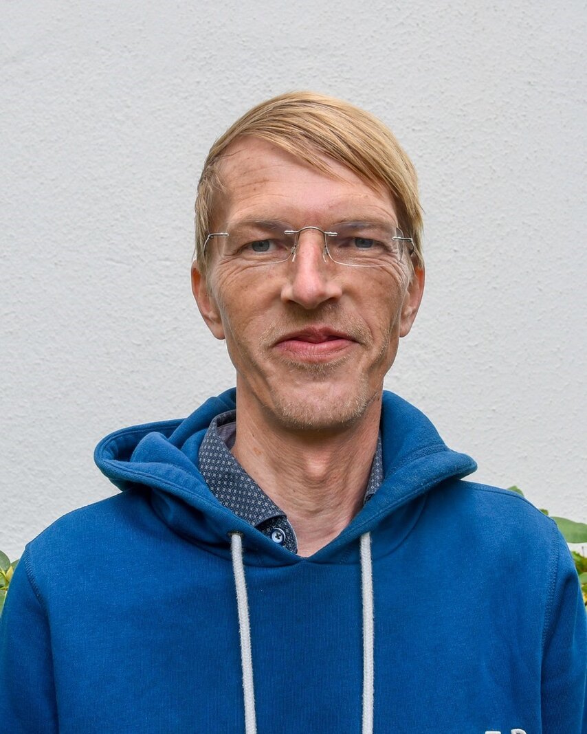 Person mit Brille und blauem Hoodie lächelt vor neutralem Hintergrund.
