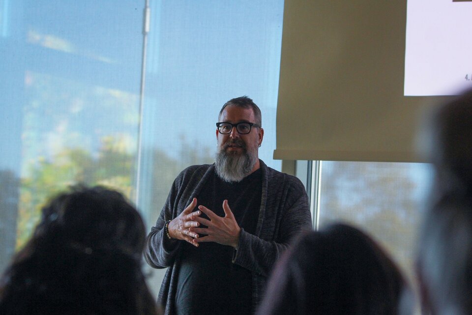Person mit Bart und Brille hält Vortrag vor einer Gruppe in einem Raum mit Fenster und Projektionsleinwand.
