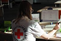 Person sitzt mit "Deutsches Rotes Kreuz"-Shirt an einem Schreibtisch vor einem Bildschirm.