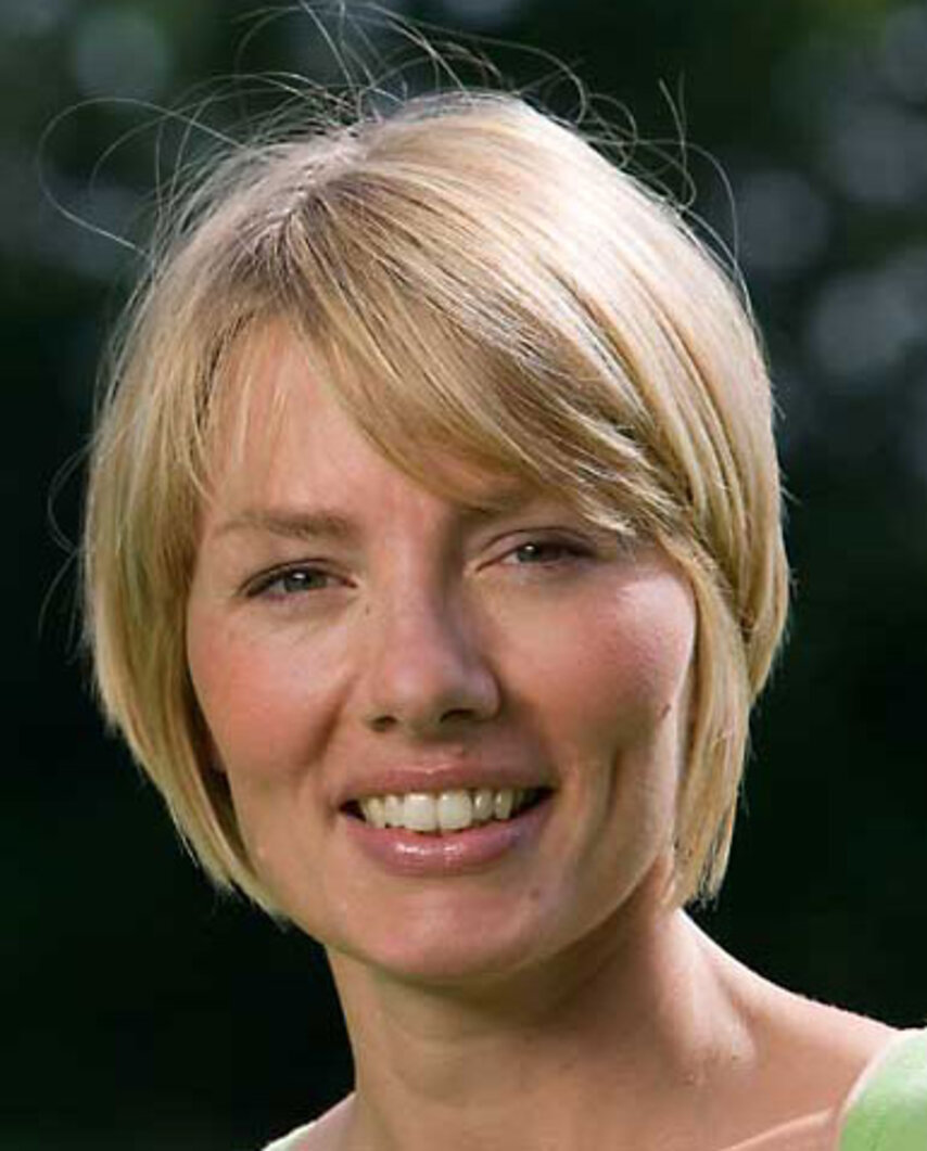 Frau mit kurzem blondem Haar lächelt im Freien, unscharfer grüner Hintergrund.