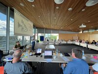 Konferenzraum mit U-förmigem Tisch, Präsentation am Bildschirm, Menschen sitzen und hören einem Sprecher zu.