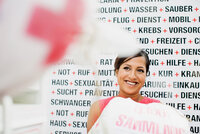 Person sitzt vor Wand mit Texten zu Gesundheitsthemen wie 'Prävention', 'Wasser', 'Mobil', 'Krebs', 'Freizeit', 'Sexualität' und 'Schwangerschaft'