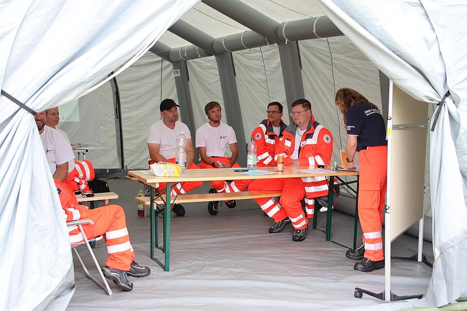 Menschen in orangefarbener Einsatzkleidung besprechen sich in einem Zelt um einen Tisch.