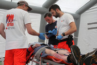 Drei Einsatzkräfte der Medical Task Force in weißem T-Shirt und oranger Hose versorgen liegende Person mit Verletzungen an den Beinen in einem Zelt