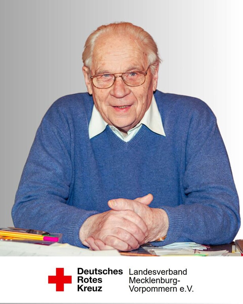 Älterer Mann in blauem Pullover sitzt an Tisch. Deutsches Rotes Kreuz-Logo unten rechts.