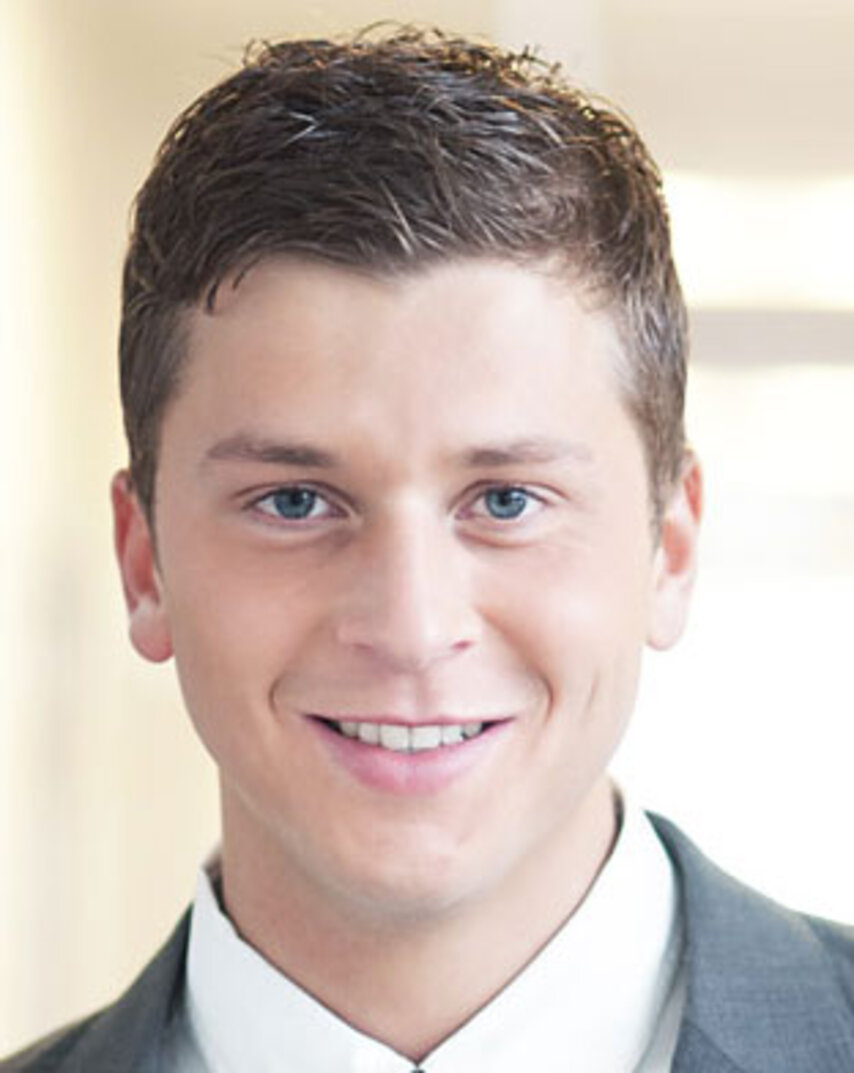 Lächelnder Mann mit kurzen, dunklen Haaren in weißem Hemd und grauem Anzug.