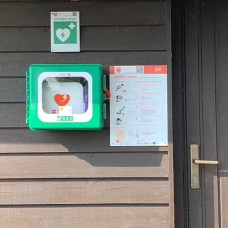 Defibrillator an Holzwand, daneben Anleitung.
