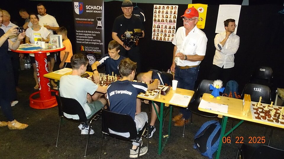 Jugendliche spielen Schach an Tischen, von Erwachsenen umgeben. Ein Kameramann filmt. Schach-Dekor an der Wand.