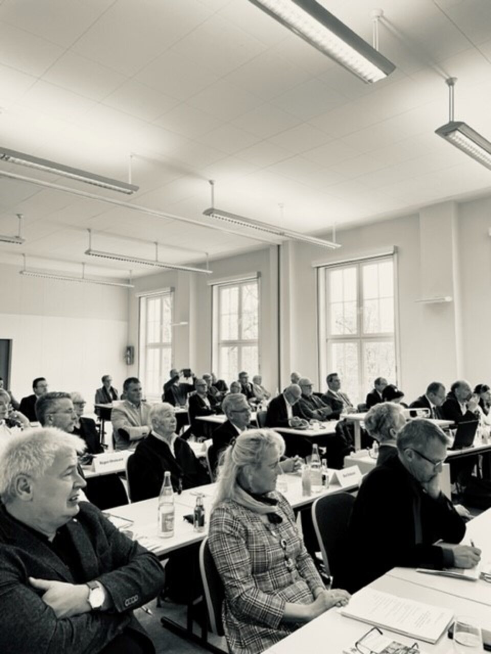 Personen in einem Konferenzraum an Tischen sitzend, notieren oder lesen Unterlagen.