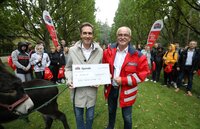 Zwei Männer halten einen Scheck, umgeben von Menschen im Park, Esel im Vordergrund.