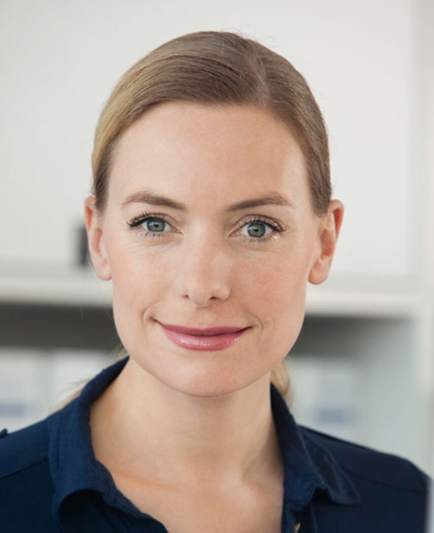 Porträt einer lächelnden Frau mit blonden Haaren, im blauen Hemd, vor unscharfen Regalen im Hintergrund.