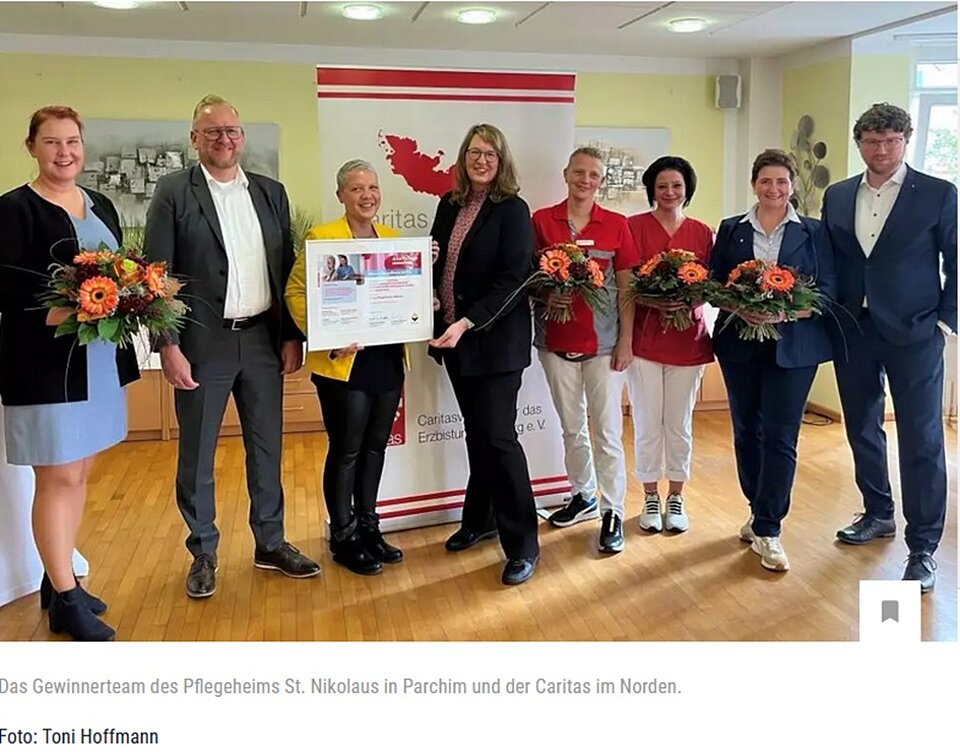 Gruppe von sieben Menschen, einige mit Blumen, lächeln vor einem Caritas-Banner. Eine hält eine Auszeichnung.