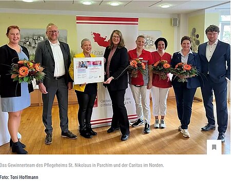 Gruppe von sieben Menschen, einige mit Blumen, lächeln vor einem Caritas-Banner. Eine hält eine Auszeichnung.