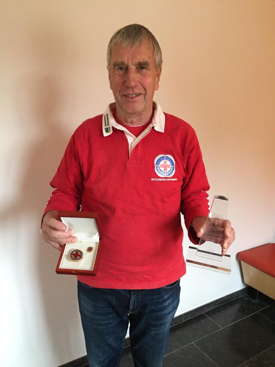 Person in rotem Pullover hält Medaille in Schachtel und transparenten Pokal.