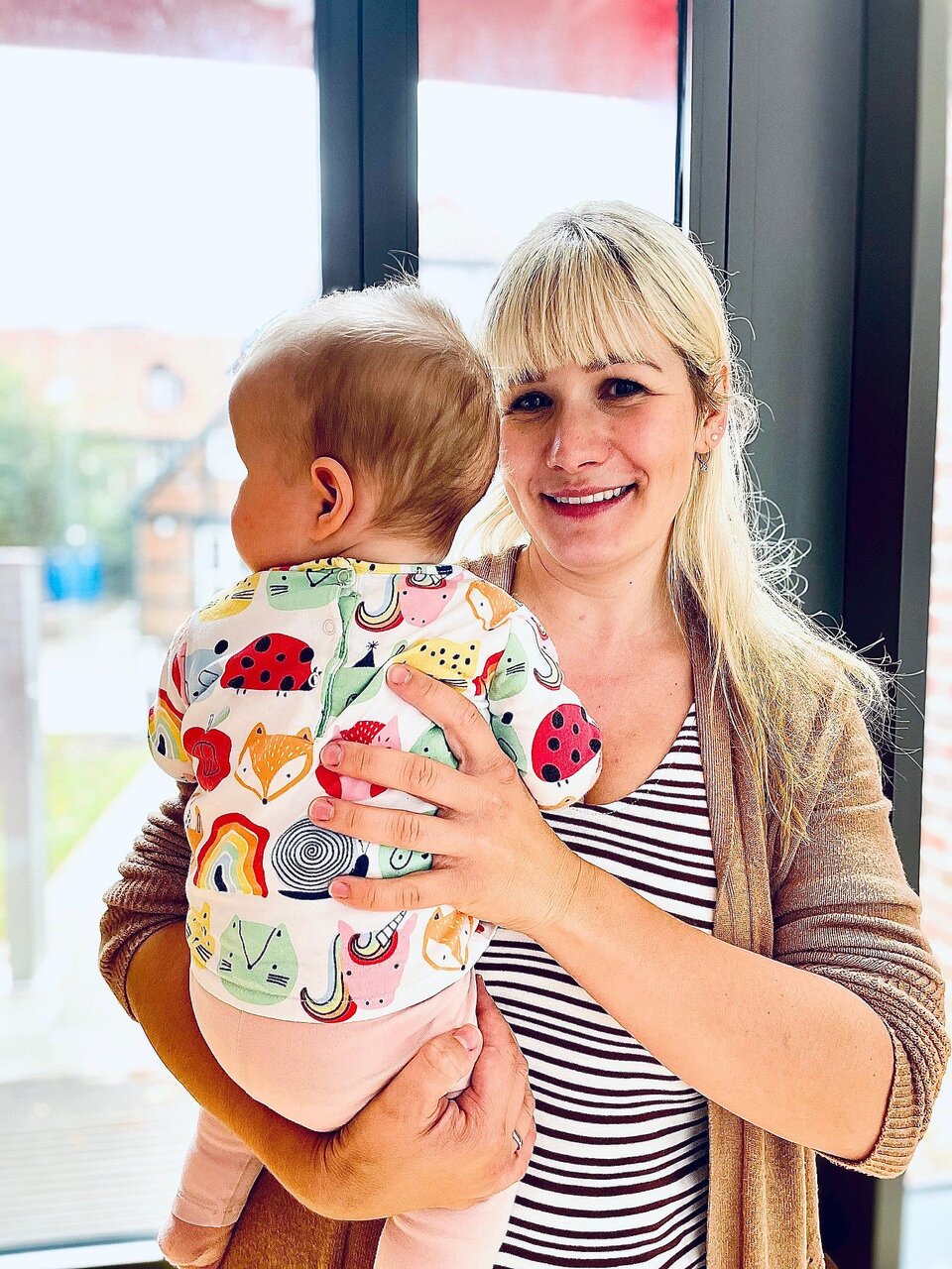 Frau hält Baby im Arm, im Hintergrund große Fenster. Baby trägt bunte Kleidung mit Tiermotiven.
