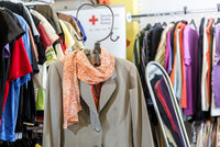 Beigefarbener Anzug mit orangefarbenem Schal, umgeben von bunten Kleidungsstücken in einem Laden.