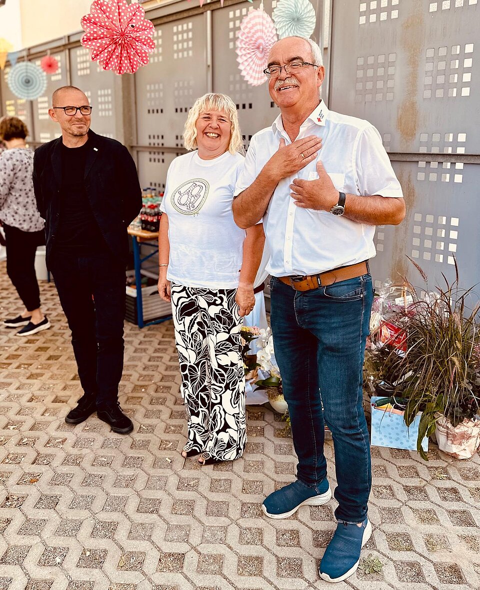 Drei lächelnde Personen stehen auf gepflastertem Boden, bunte Dekorationen hängen im Hintergrund.