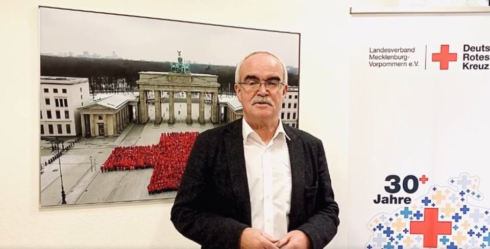 Mann vor DRK-Poster, daneben Bild vom Brandenburger Tor mit Menschenformation in Form eines roten Kreuzes.