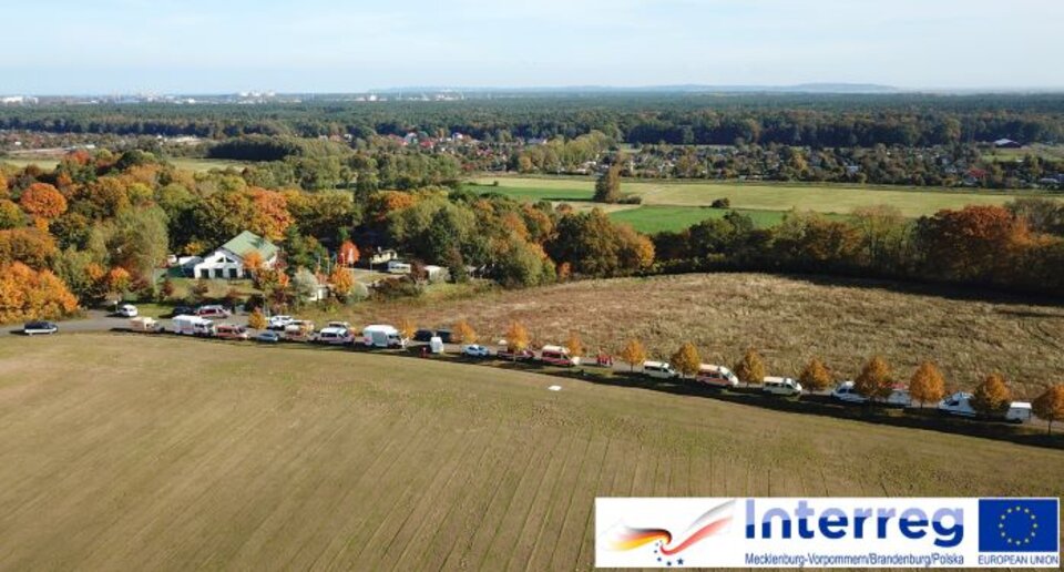 Luftaufnahme: Feld mit parkenden Autos entlang einer Straße, herbstliche Bäume, dahinter Häuser und Wälder.