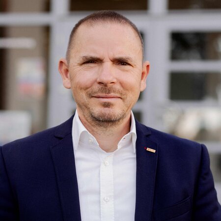 Mann in blauem Anzug und weißem Hemd steht draußen vor einem Gebäude mit verglaster Tür.