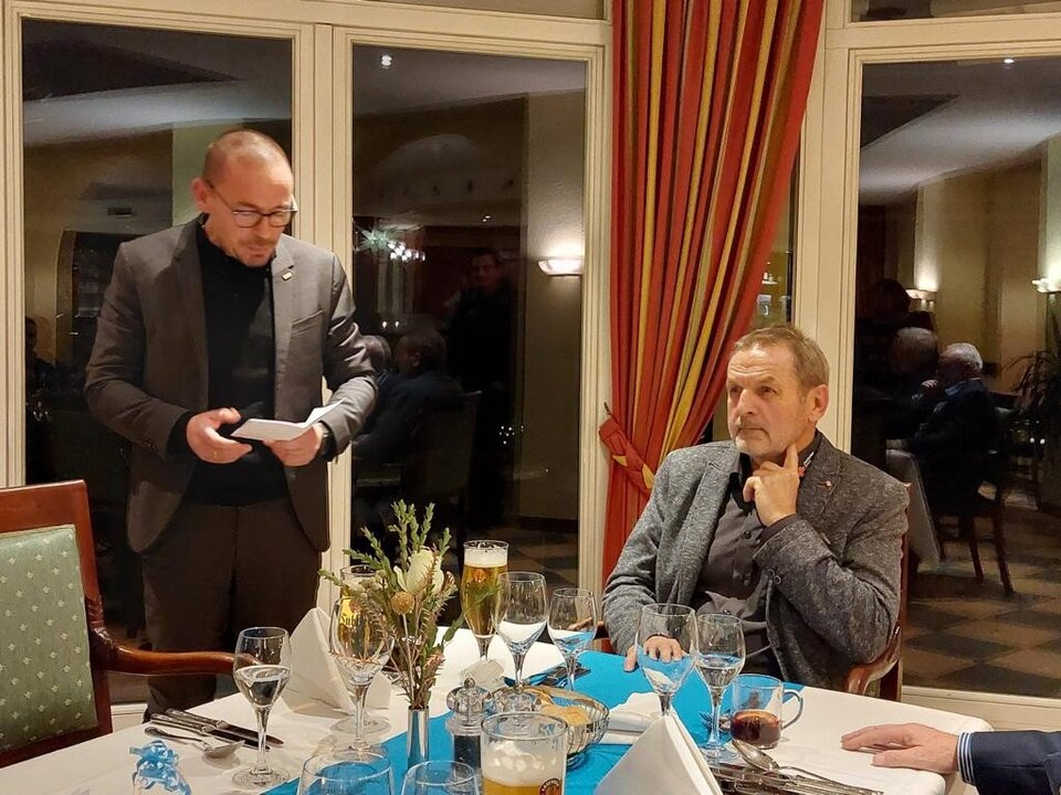 Mann hält Rede neben sitzendem Mann an gedecktem Tisch in Restaurant mit rot-gelben Vorhängen.