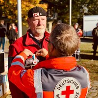 Rotes Kreuz Mitglied verleiht einem anderen Mann eine Auszeichnung im Freien. Beide tragen Uniformen des Roten Kreuzes.