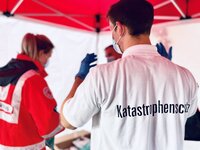 Person mit weißem Hemd und Aufschrift 'Katastrophenschutz' auf dem Rücken, trägt Mundschutz und blaue Handschuhe, spricht mit einer weiteren Person in roter Einsatzkleidung unter einem roten Zelt