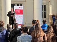 Mann spricht zu Gruppe, neben Banner des Deutschen Roten Kreuzes zur Migrationsberatung für erwachsene Zuwanderer.