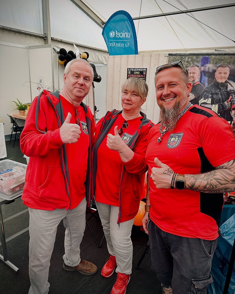 Drei Personen in roten T-Shirts und Kapuzenjacken, zeigen Daumen hoch, im Zelt mit UNICEF-Logo im Hintergrund.