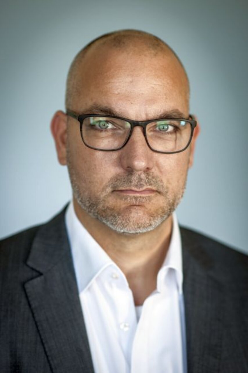 Mann mit Brille, Glatze und Bart in Anzug und weißem Hemd vor grauem Hintergrund.